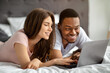 © Prostock-studio - Interracial loving couple spending weekend together at home, using laptop while lying on bed