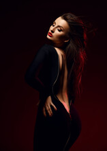 Sexy Brunette Woman Vamp Stands Sideways In Black Tight Unzipped Dress Demonstrating Her Naked Back, Waistline And Butt Over Black Background . Fashion, Vogue, Sexy Stylish Look For Woman Concept