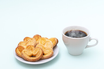 White cup of steaming hot black coffee or hot chocolate and freshly cooked heart shaped cookies on a light blue background, copy space. Morning breakfast for loved ones