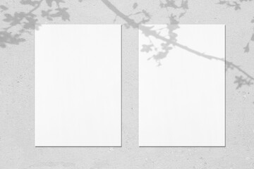 Two empty white vertical rectangle poster mockups with soft shadows of blooming tree leaves and flowers on neutral light grey concrete wall background. Flat lay, top view