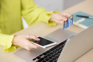 Closeup young asian business woman using smart phone and holding credit card while online shopping and payment with laptop computer on desk at home, female holding debit card, communication concept.