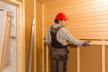 Sauna construction, finishing. The man is screwing a wooden bench to the wall.