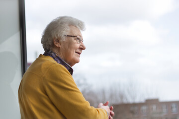 Horizontal portrait of an old senior woman, 80s adult, grandmother, standing on her balcony, leaning on the balustrade, smiling, thinking and looking in the distance. Copy space.