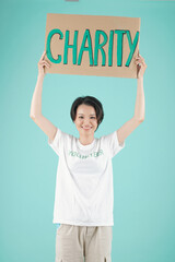 Wall Mural - Smiling young Asian volunteer standing with charity placard, isolated on light blue