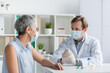 © LIGHTFIELD STUDIOS - Doctor in medical mask sitting near mature patient and vaccine with pills during consultation