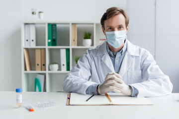 Wall Mural - Doctor in medical mask and latex gloves looking at camera near notebook, vaccine and syringes on blurred foreground