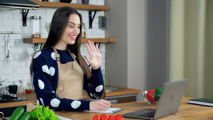 Smiling woman chef teacher in beige apron greets housewife student teaches online remote video call conference webcam chat laptop computer, distance education learning cook course in home kitchen