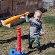 A toddler boy watches his baseball fly away after knocking it off the tee with a plastic bat on a sunny spring day. 