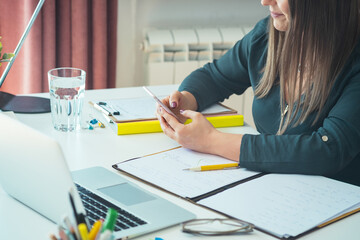 Wall Mural - Cropped photo of unrecognizable female teacher sitting at home office desk and holding mobile phone. Young professor or student having break with online class and using smartphone to send text message