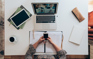 Wall Mural - Businesswoman using laptop and phone for video conference call meeting with colleagues. Top view of home office desk with business papers, tablet, coffee, computer and female hands using mobile phone