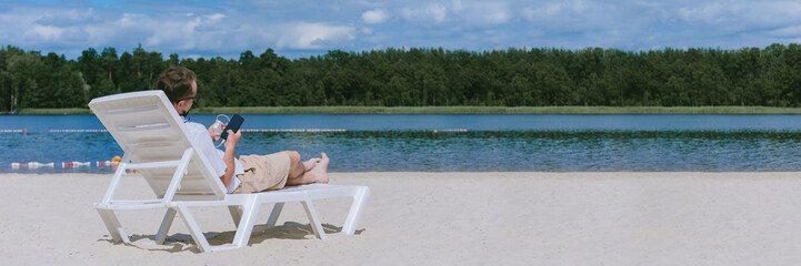 Wall Mural - Banner, a man lies on a sun lounger and charges a smartphone from a Power Bank on the beach. Against the backdrop of sand, water and forest.