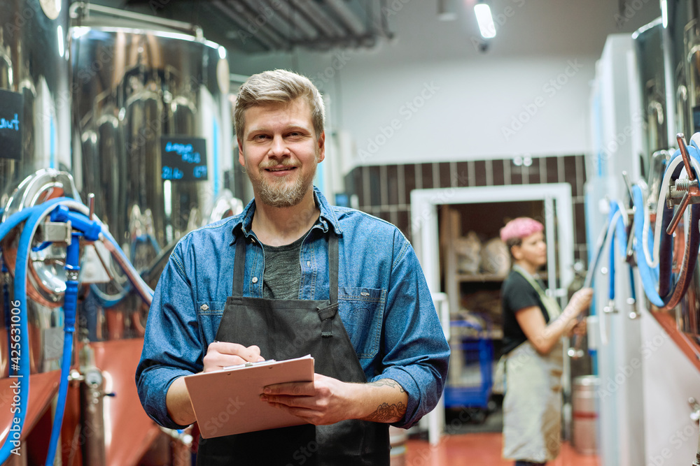 Young bearded male worker of large contemporary brewery making notes while standing against female brewer using processing machine - obrazy, fototapety, plakaty 
