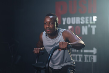 Wall Mural - African ethnicity man listening music while using spinning bike on cardio machine in fitness gym