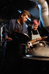 Sticker - Young woman with tablet scrolling through list of beer processing stages while standing next to her male colleague opening lid of cistern