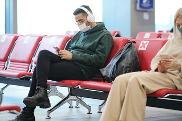 Wall Mural - Young man with headphones listening to music and scrolling in smartphone while sitting in lobby of modern airport and waiting for flight