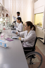 Canvas Print - Disabled scientist sitting at the table and working with samples in test tubes with her colleagues in the background