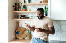 Focused Young African American Stylish Bearded Man Standing At Kitchen, Holding A Glass Of Pure Water And Smartphone In Hands, Browsing Internet, News Or Chatting With Friends