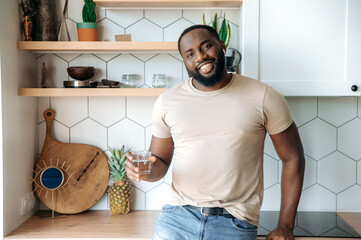 A young handsome healthy African American bearded guy stands at kitchen, in casual clothes, with a glass of clean water in his hand, looks at the camera, smiling friendly, leads healthy lifestyle