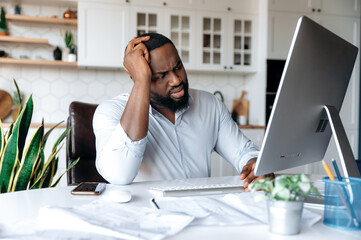 Wall Mural - Upset sad young african american man, manager, broker or freelancer, working at home at the computer, experiencing stress at work, holding his head with his hand, looking sadly at the screen