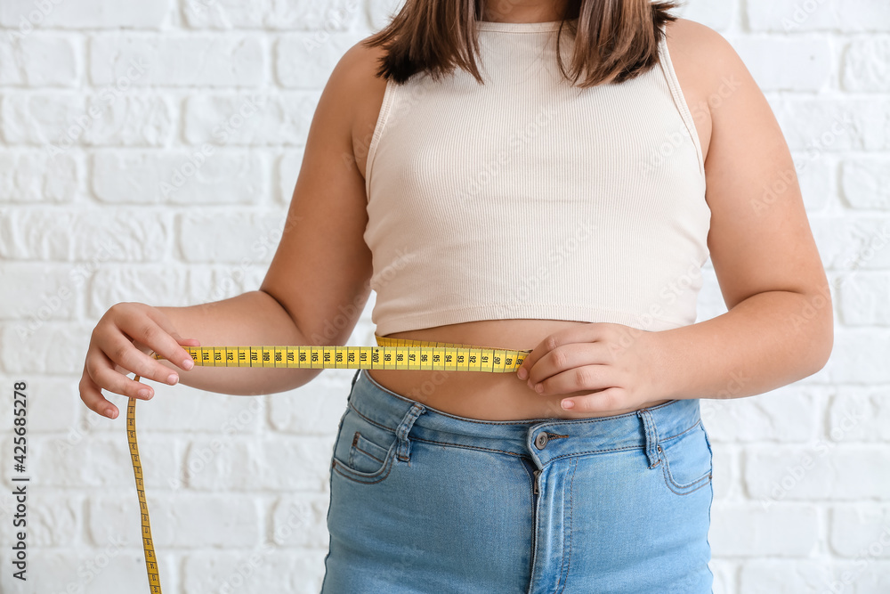 Overweight girl measuring her waist on white brick background
