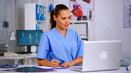 Wall Mural - Doctor assistant in uniform working on laptop in modern hospital office. Therapist writing list of consulted patients online, making research, noting, taking useful information on clipboard