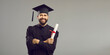 © Studio Romantic - Portrait of a happy student in an academic dress and hat having a university or academy certificate. Man with folded arms smiles and looks at the camera on a gray background. Graduate concept. Banner.