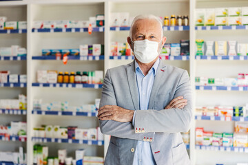 Wall Mural - Senior doctor with his arms crossed is standing in a pharmacy. A portrait of a man working in a pharmacy in an elegant suit and with a protective mask on his face. Protection against corona virus