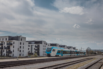 Wall Mural - Modern white and blue passenger train on a station in front of a modern housing or residental district. City blocks and train.