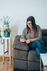 Wall Mural - Cheerful woman sitting on sofa and using mobile phone at home.
Smiling businesswoman working at home, calling or typing message on smartphone next to the plants in flowerpot in living room