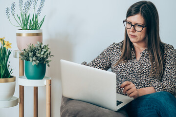 Wall Mural - Business woman sitting on couch, talking at online video conference with clients or partners on laptop, working at home next to the potted flowers in living room
