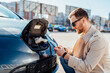 © Andrii  - Casual man with smartphone near electric car waiting for the finish of the battery charging process
