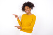 © Roquillo - young beautiful African American woman wearing yellow sweater against white wall points aside with  surprised expression with mouth opened, shows something amazing. Advertisement concept.