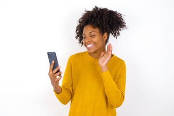Portrait of happy friendly young beautiful African American woman wearing yellow sweater against white wall taking selfie and waving hand, communicating on video call, online chatting.