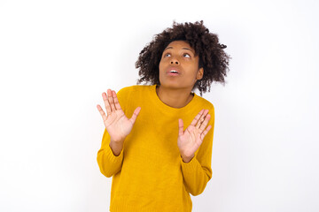 Wall Mural - young beautiful African American woman wearing yellow sweater against white wall keeps palms forward and looks with fright above on ceiling tries to defense herself from invisible danger opens mouth.