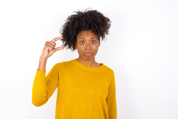 Wall Mural - young beautiful African American woman wearing yellow sweater against white wall purses lip and gestures with hand, shows something very little.