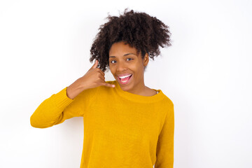Wall Mural - young beautiful African American woman wearing yellow sweater against white wall makes phone gesture, says call me back again, has glad expression.