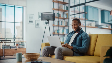 Wall Mural - Handsome Black African American Man Working on Laptop Computer while Sitting on a Sofa in Cozy Living Room. Freelancer Working From Home. Browsing Internet, Using Social Networks, Having Fun in Flat.