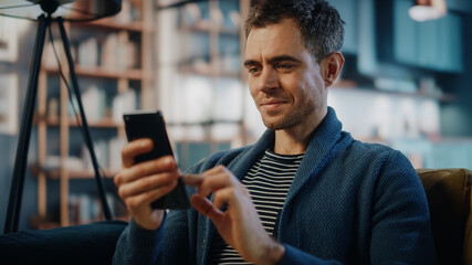 Excited Caucasian Man Using Smartphone while Resting on a Sofa in Stylish Living Room. Happy Man Smiling at Home and Chatting to Colleagues and Clients Over the Internet. Using Social Networks.