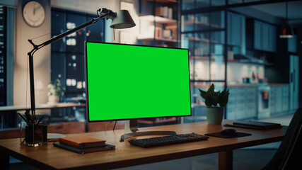 Desktop Computer with Mock-up Green Screen Dsiplay Standing on the Wooden Desk in the Creative Cozy Living Room. In the Background Warm Evening Lighting and Open Space Studio.