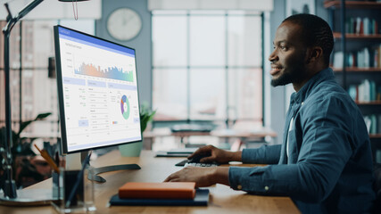Handsome Black African American Specialist Working on Desktop Computer in Creative Home Living Room. Freelance Male is Doing Market Analysis and Creates Report with Charts for Clients and Employer.