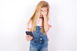 © Roquillo - beautiful caucasian little girl wearing denim jeans overall over white background looking at smart phone feeling sad holding hand on face.