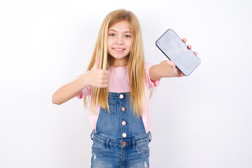 Wall Mural - Portrait beautiful caucasian little girl wearing denim jeans overall over white background holding in hands cell showing giving black screen thumb up