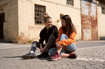 Two teenage skater girls are hanging out in the neighborhood, chatting and smiling.