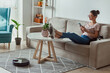 © perfectlab - Robotic vacuum cleaner cleaning carpet, woman remote control mobile phone and enjoy rest, sitting on sofa at home