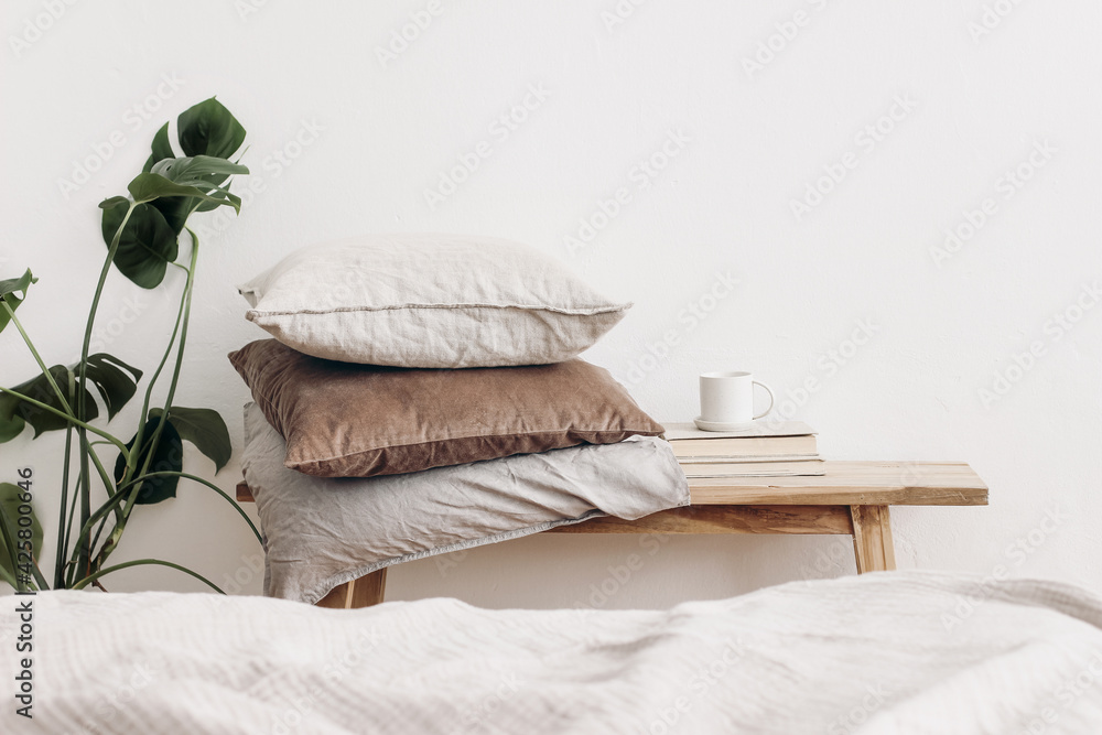 Trendy Scandinavian interior still life. Velvet and linen cushions on vintage wooden bench, table. Cup of coffee on pile of books and monstera potted plant. White wall background. Blurred bed - obrazy, fototapety, plakaty 