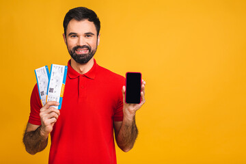 Wall Mural - Portrait of stylish handsome bearded young man, isolated over yellow background. Man smiling, looking at camera and holding tickets. Showing mobile phone screen.