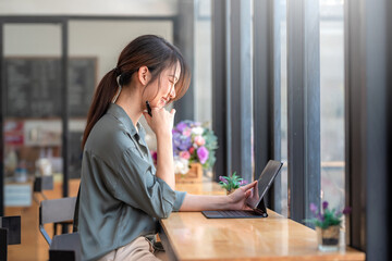 Side view Asian woman sitting by the window of a happy cafe using a tablet.