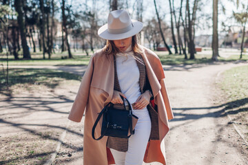 Wall Mural - Stylish woman wears hat holding handbag walking in park. Spring female clothes and accessories. Fashion