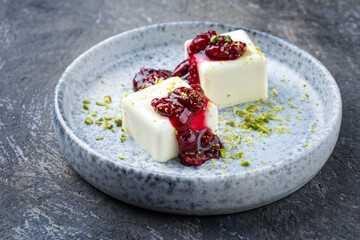 Modern style traditional blancmange almond pudding with sour cherry coulis and pistachios served as close-up in a Nordic design plate on gray background