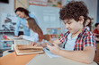 © Ivan - Young boy playing smartphone whine lesson. Elementary school kids sitting on desks and reading books in classroom